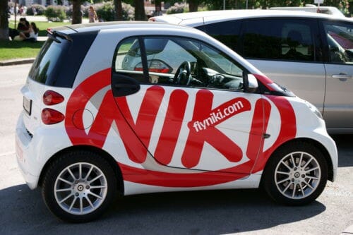 "Vienna, Austria - May 1, 2012: Small Smart car with logo of Niki airline. Niki, also known as flyNiki was founded by ex-F1 Grand Prix driver Niki Lauda in 2003. It is owned by Air Berlin since November 2011. People in background enjoying in the park."